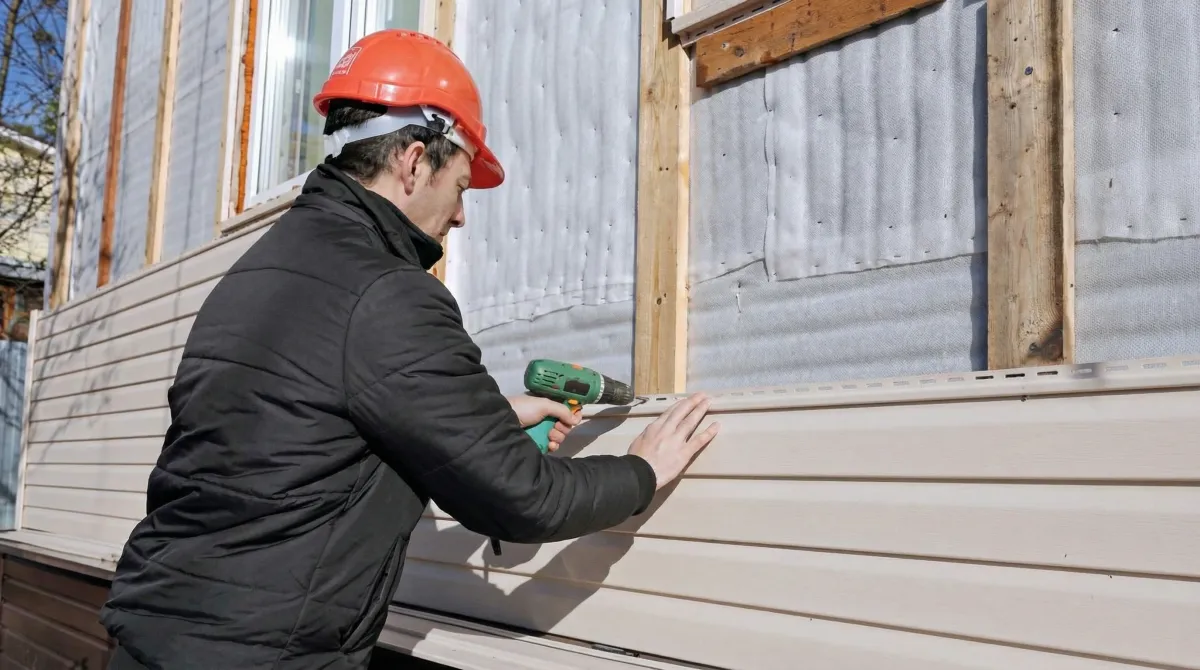 Siding replacement professional using a power drill to install new tan vinyl siding on a home exterior in Jersey City, NJ
